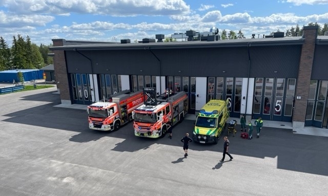 Harmaa pelastusasema, jonka pihassa kaksi paloautoa, ambulanssi sekä henkilökuntaa. Aurinkoinen sää.